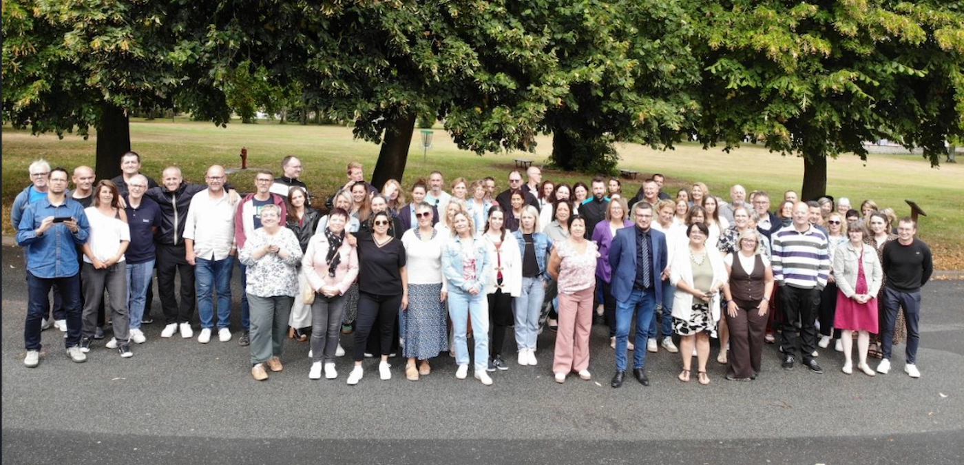 Un grand groupe d'adultes posant ensemble pour une photo d'équipe en extérieur, sur une cour goudronnée avec de grands arbres et un espace vert en arrière-plan